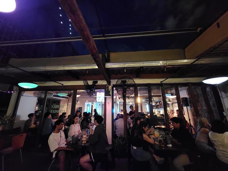 Groups of drinkers at dark tables on a covered rooftop terrace, with a lit indoor bar room visible through glass doors behind them.