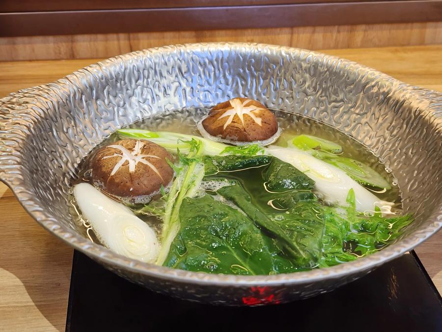 Shiitake mushrooms, leek, mizuna and spinach simmering in clear broth in a hammered metal pot on an induction hob.