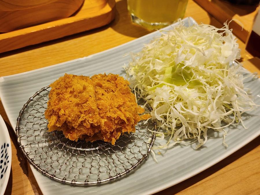 A single bite-size pork fillet cutlet on a round wire rack beside a pile of shredded cabbage on a pale blue plate.