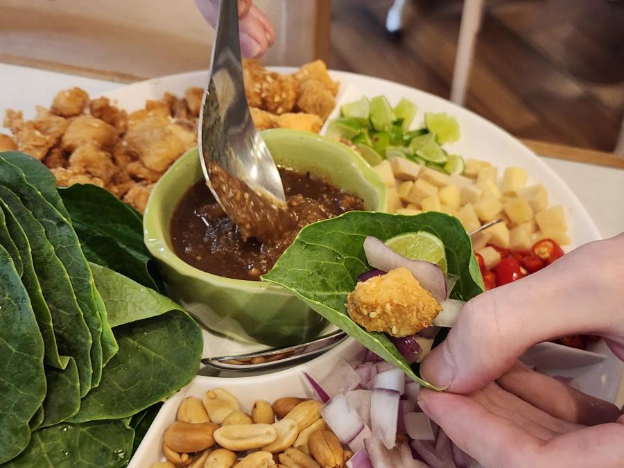 A hand holding a leaf folded around a piece of fried sea bass, shallot and lime, with a spoon pouring dark sauce over it.