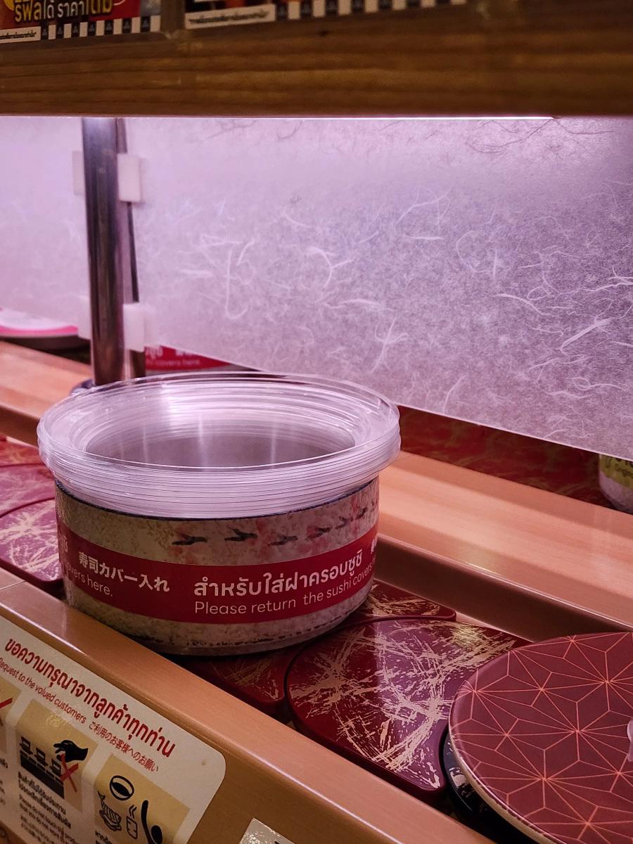 A speckled ceramic bin stacked with clear plastic sushi covers on the counter, labelled in Japanese, Thai and English.