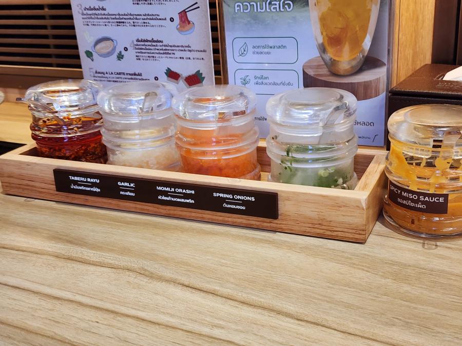 A wooden tray of five lidded glass jars labelled taberu rayu, garlic, momiji oroshi and spring onions, with a jar of spicy miso sauce beside it.