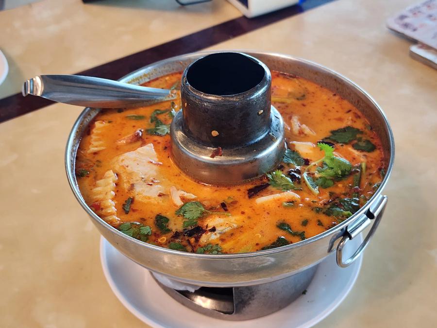 A steel hot pot of orange tom yum soup with pieces of squid and fish, coriander leaves and a ladle.