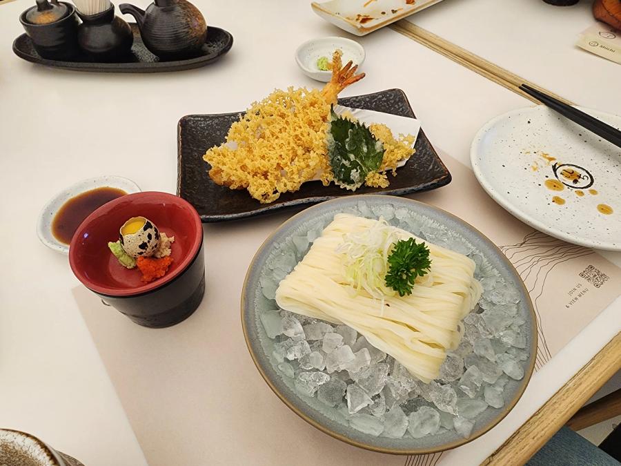 Cold inaniwa udon on crushed ice with spring onion and parsley, a red cup holding a quail egg, wasabi and tobiko, a dish of dipping sauce and a plate of prawn tempura with a shiso leaf.