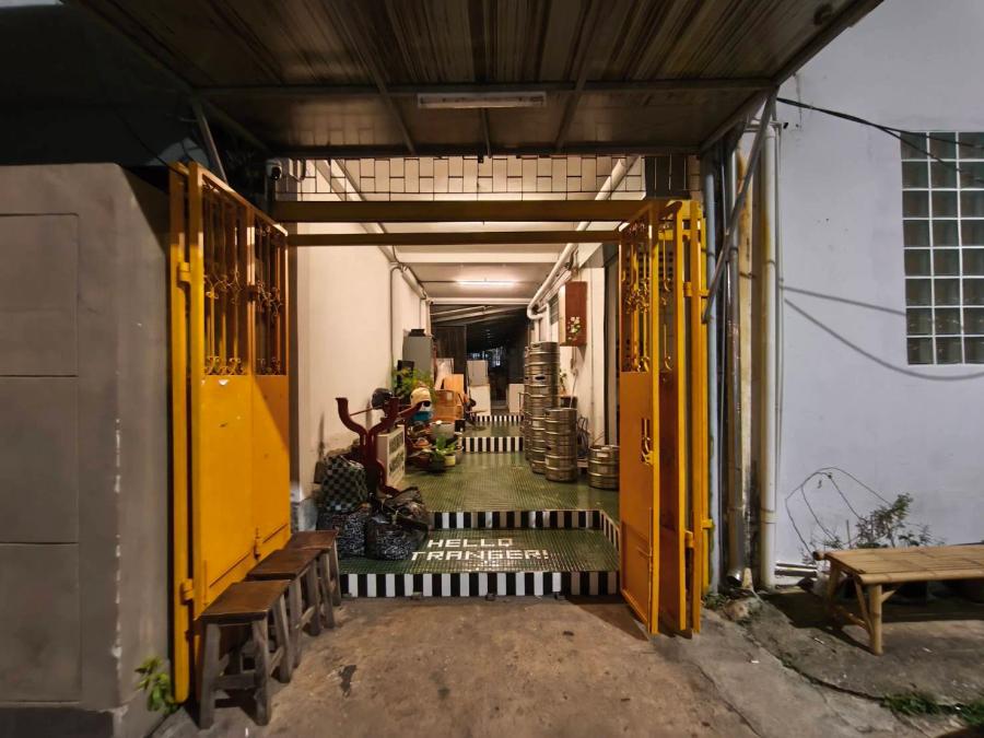 A yellow folding gate open on a shophouse passage, with Hello Stranger spelled in tiles on a black and white striped step and beer kegs stacked inside.