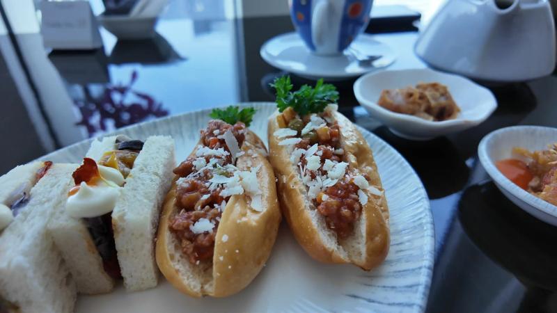 A plate of finger sandwiches and two small hot dog buns topped with minced sauce and grated cheese on a dark glass table.