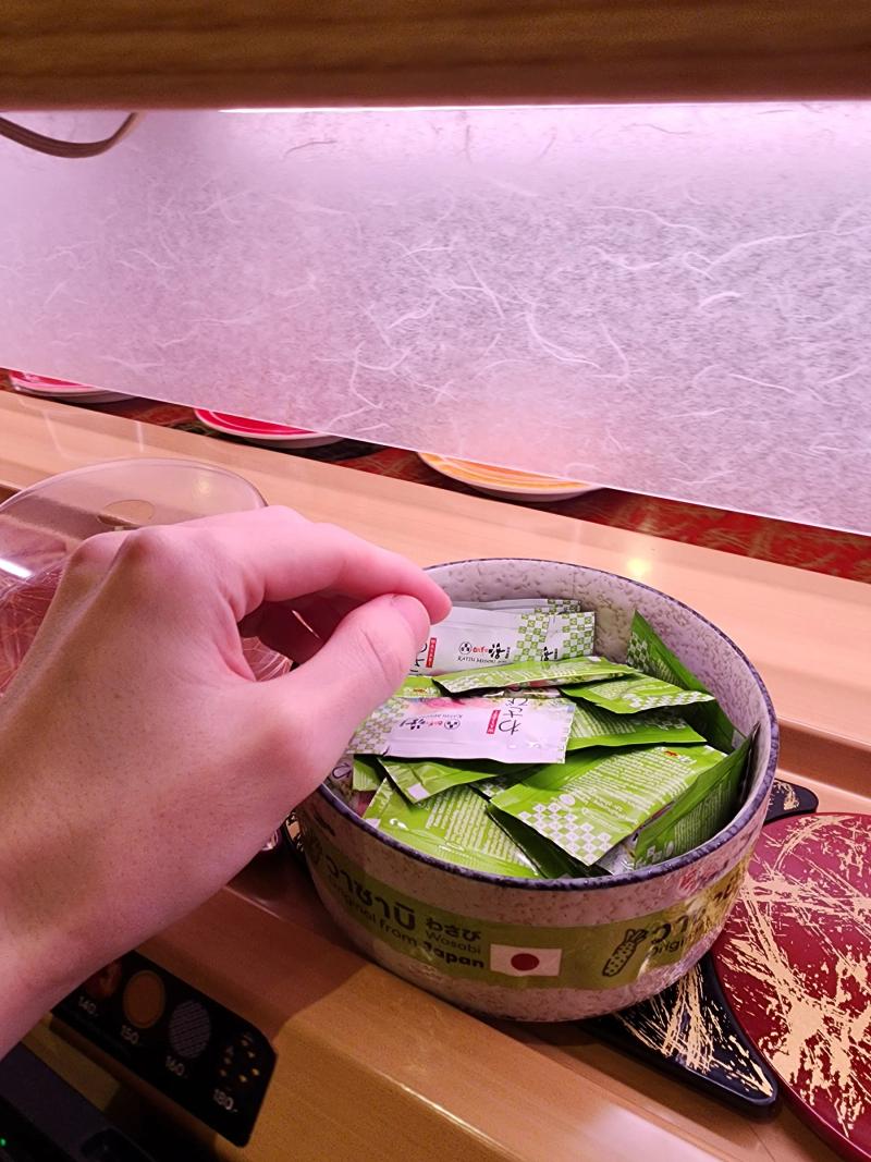 A hand taking a wasabi sachet from a speckled bowl on the counter, with plates passing on the belt behind a paper screen.