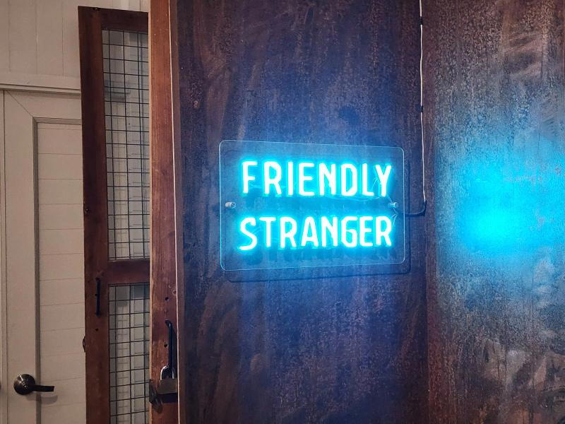 A blue neon sign reading Friendly Stranger mounted on a dark timber panel beside a white door.