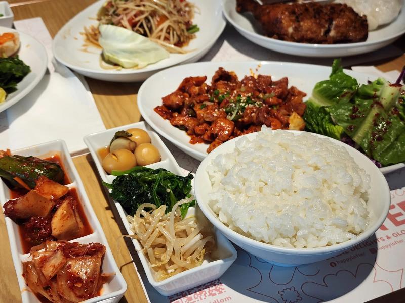 A Korean spicy pork set with rice and two trays of side dishes, with papaya salad and fried chicken behind.