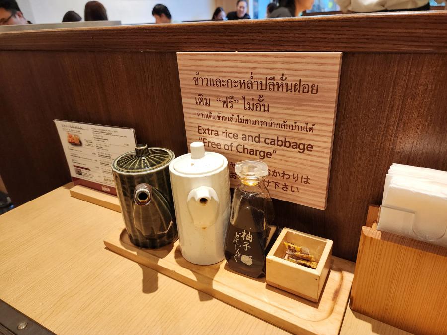 Two ceramic sauce pots, a bottle of yuzu dressing and a box of sesame sachets on a wooden tray under a wooden sign reading extra rice and cabbage free of charge in Thai, English and Japanese.