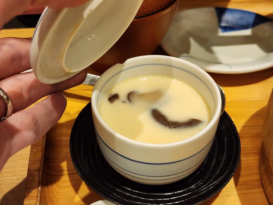 A hand lifting the lid from a cup of chawanmushi steamed egg with a slice of mushroom on top, on a black saucer.