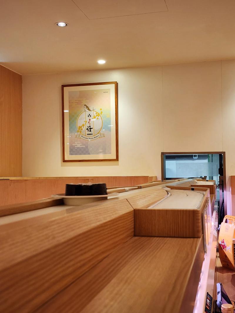 A framed Welcome Katsu Midori print with a fish and yellow blossom on a cream wall, above a wooden counter with a plate of maki on the belt.