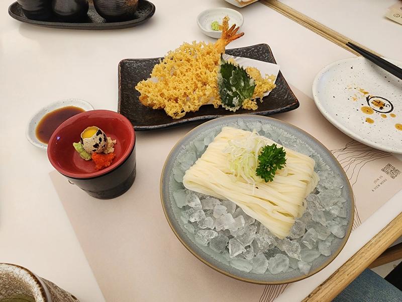 Cold inaniwa udon on crushed ice, a red cup with a quail egg, wasabi and tobiko, a dish of dipping sauce and a prawn tempura with a shiso leaf, seen from above.