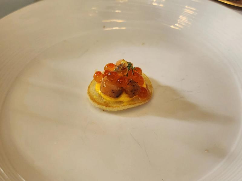 A small blini topped with yellow cream, cubes of cured salmon, orange salmon roe and a sprig of dill.
