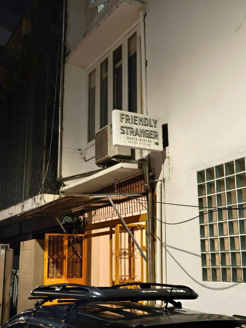 A white lightbox sign reading Friendly Stranger Bar and Bistro, Rooftop 4th Floor, fixed to a white shophouse above a yellow folding gate.