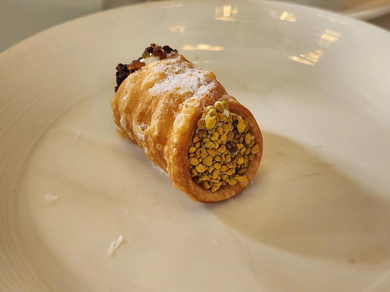 A small cannoli dusted with icing sugar, one end filled with yellow bee pollen and the other with dark chopped chocolate.
