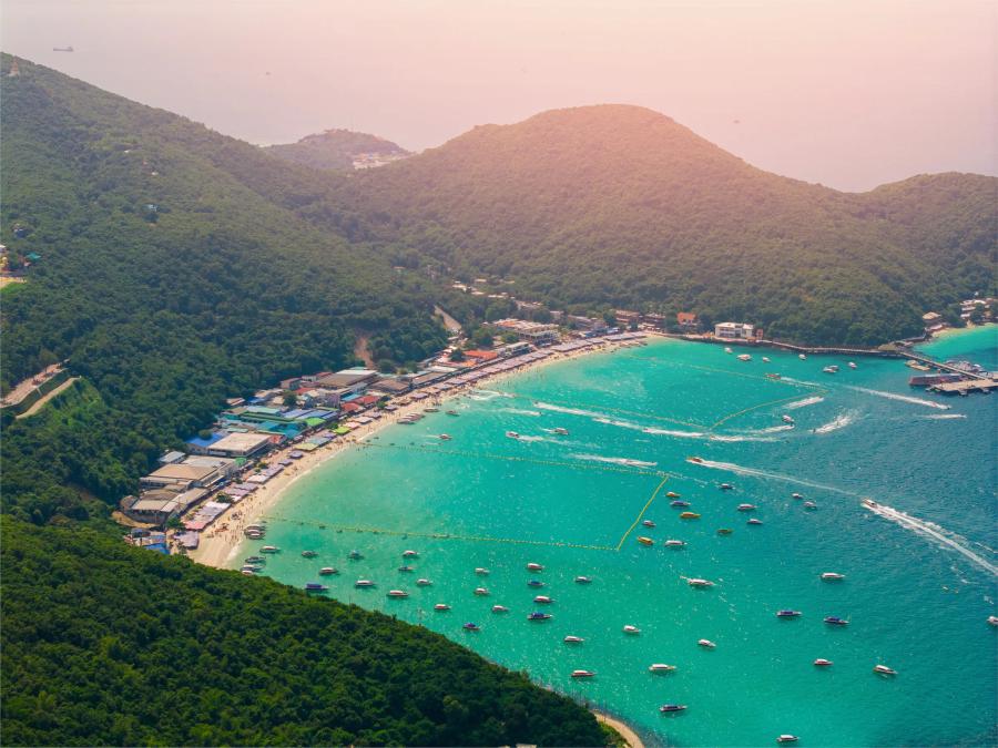 An aerial view of Tawaen Beach on Koh Larn, a curved bay of pale sand and turquoise water dotted with boats below green hills.