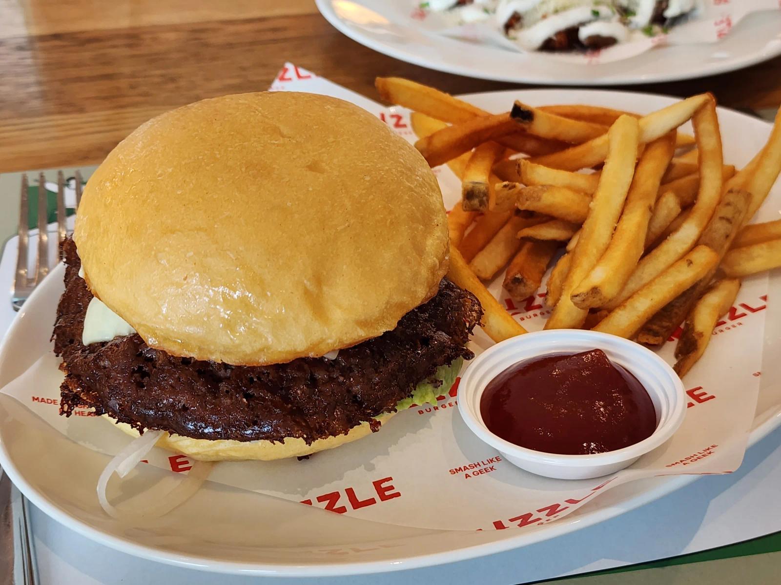 A Smizzle smash burger with a charred beef patty beside french fries and a pot of ketchup.