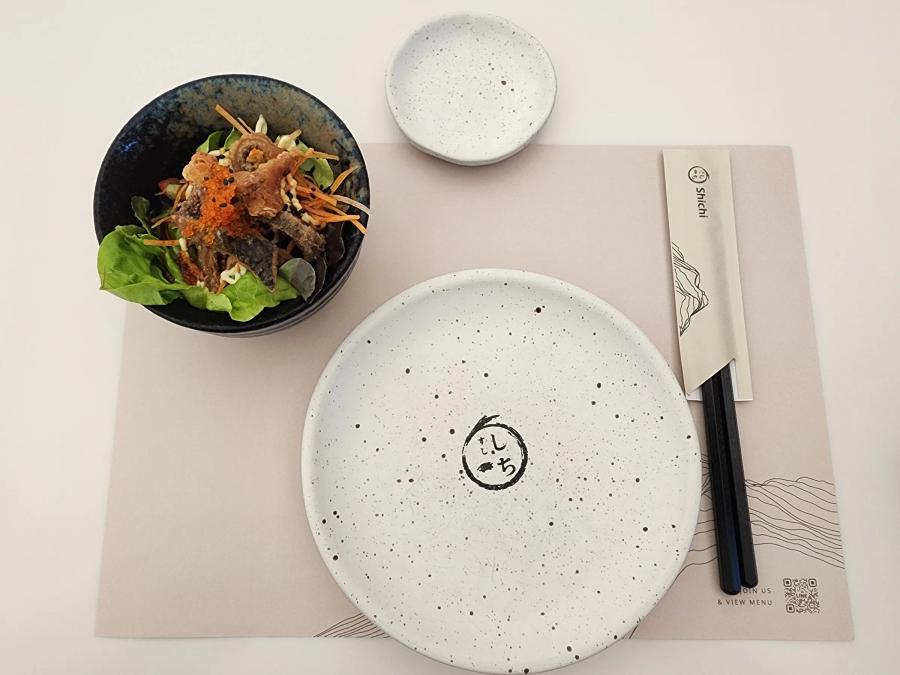 Setting from above: a white speckled plate with the restaurant logo, a small dish, chopsticks in a paper sleeve and a bowl of salad with fried silver fish and orange roe on lettuce.