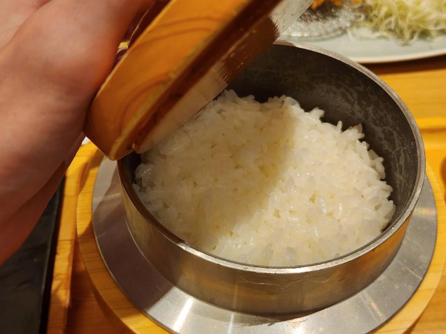A hand lifting the wooden lid from a small iron pot of freshly cooked white Japanese rice.