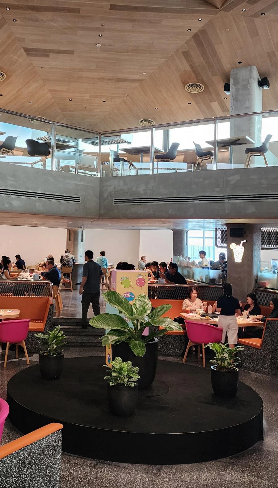 A round black platform with potted plants in the LOFTER seating hall, orange sofa booths behind it and a gallery of tables under a timber ceiling above.
