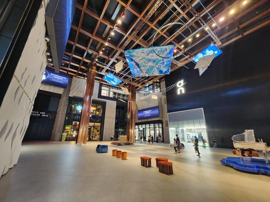 The atrium of The Storeys at One Bangkok at night, with copper columns, lit panels hanging from the ceiling, an acrylic grand piano on a blue stage and shoppers crossing the floor.