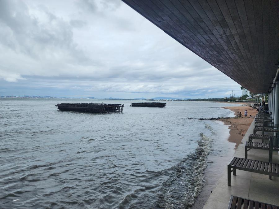 The seafront terrace at Moom Aroi with wooden benches under a deep roof, a narrow strip of sand and two timber rafts moored offshore under grey cloud.