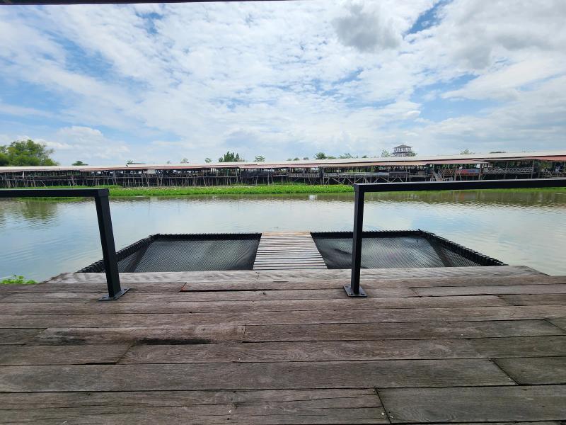An empty black net platform strung over a canal at the end of a wooden deck, with a plank walkway down the middle and a stilt market beyond.