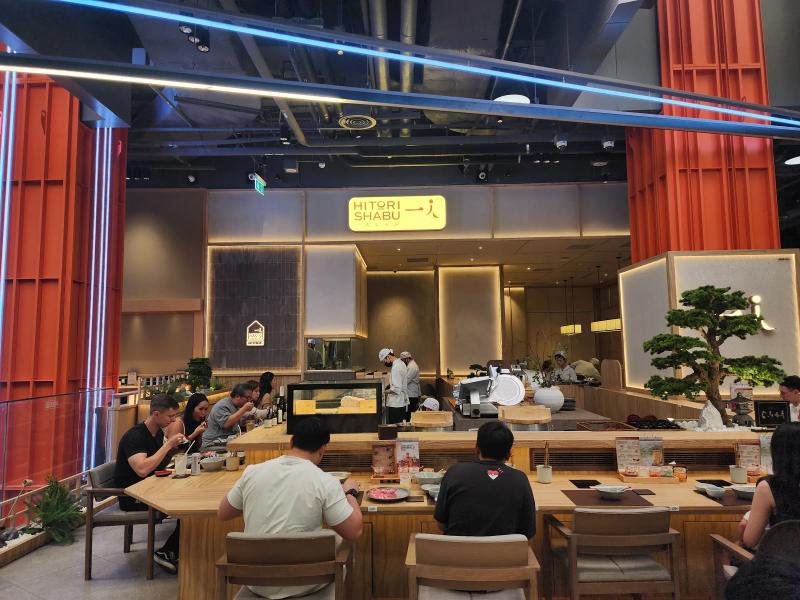 The front of Hitori Shabu One Bangkok from the left, with its lit sign, orange lattice pillars, chefs at the open kitchen and diners at the counter.