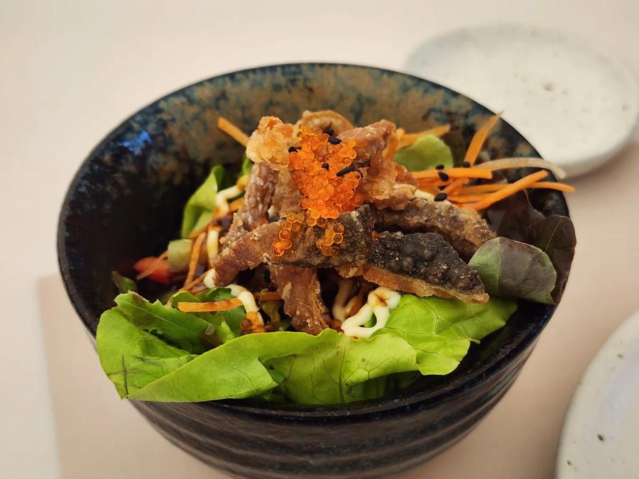 A small bowl of salad with crisp fried silver fish, orange tobiko, shredded carrot and mayonnaise on green and red lettuce.