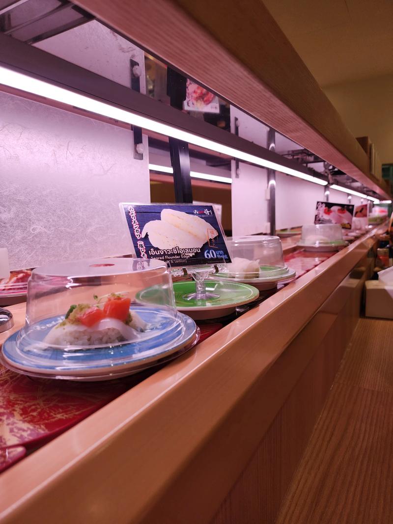 Domed sushi plates moving along the Katsu Midori belt past a card for seared flounder edge with salt and lemon at 60 baht.