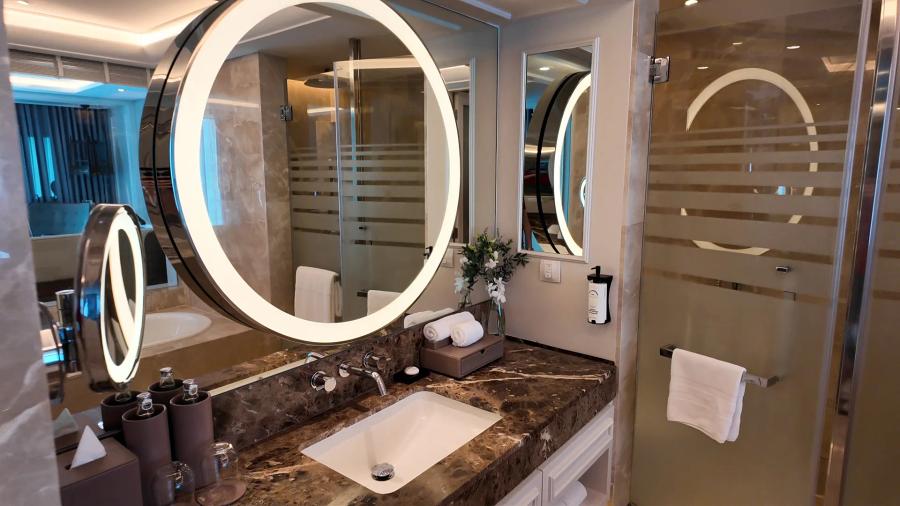 Twin basins under two ring-lit round mirrors on a dark marble counter in a hotel bathroom, with rolled towels and a leather amenity box.