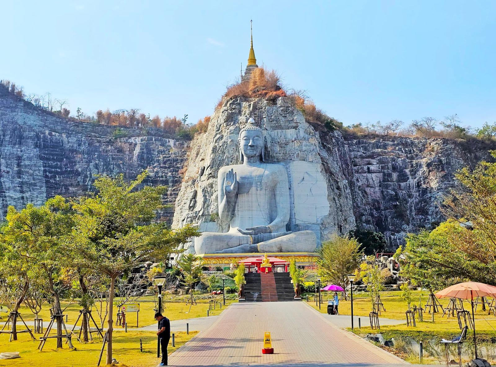A seated Buddha carved into a grey limestone cliff face at Wat Khao Tham Thiam in U Thong, Suphan Buri, with a gold chedi on the summit and a paved path leading up to it.