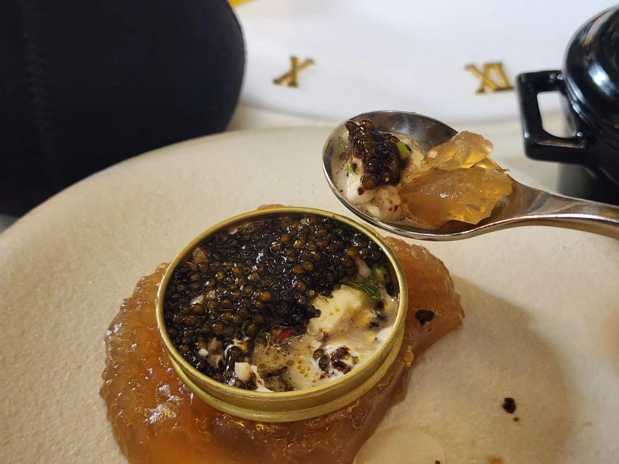 A spoon holding caviar, white scallop tartare and a piece of amber jelly above the opened gold tin.