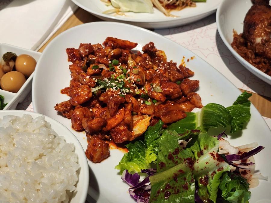 Korean spicy stir-fried pork with sesame seeds and a dressed salad, beside a bowl of rice and a dish of quail eggs.
