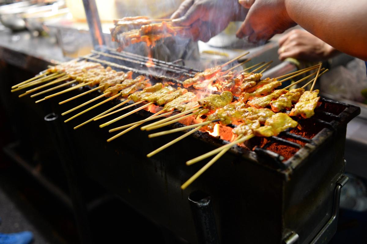 Street food vendor cooking Satay on a Bangkok pavement at dusk.