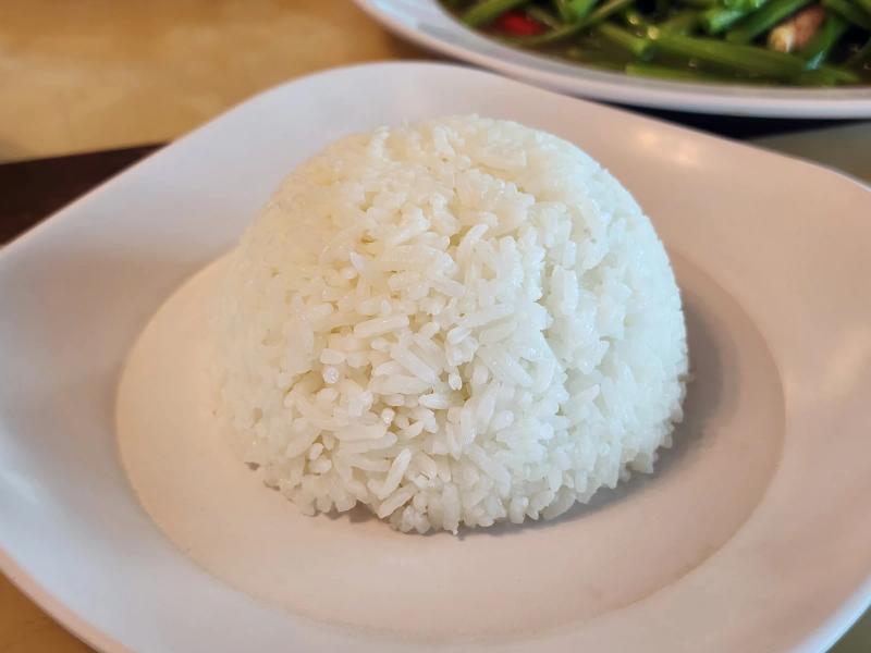 A mound of steamed jasmine rice on a white plate, with a plate of stir-fried morning glory behind.