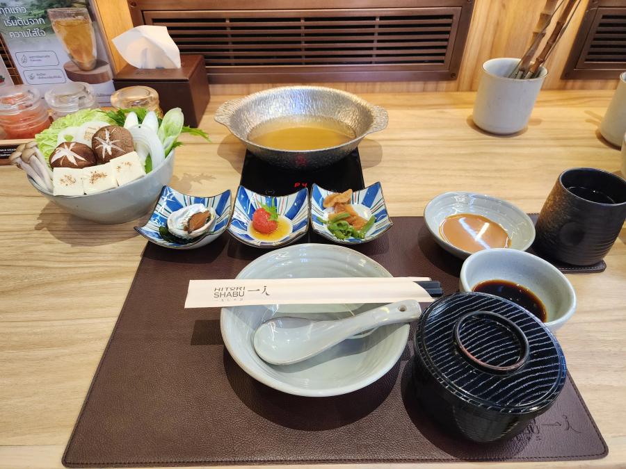 A Hitori Shabu counter setting with a bowl of vegetables, a pot of broth on an induction hob, three small starter dishes, sesame and ponzu dips, a lidded rice bowl and chopsticks on a leather mat.