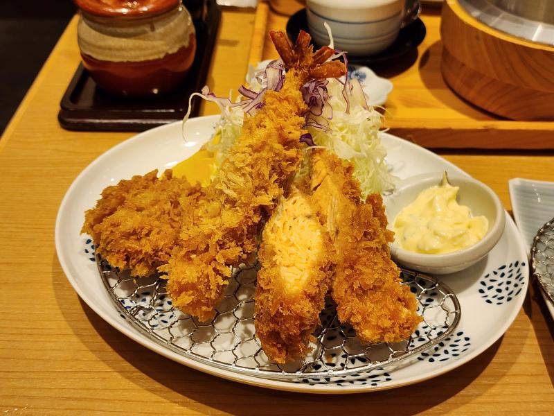 A breaded prawn, a fried salmon fillet and a bite-size pork fillet cutlet on a wire rack with cabbage and tartare sauce on a white plate.