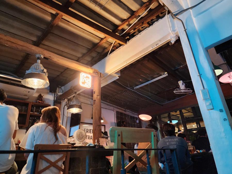 The indoor room at Friendly Stranger under a corrugated roof with timber beams, an orange FS neon sign on a post and customers seated facing the bar.