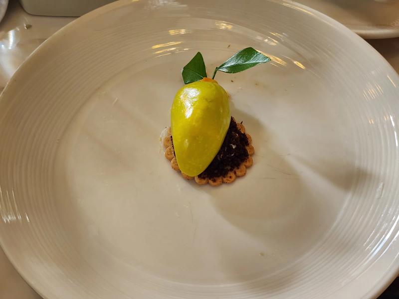 A glossy green and yellow sweet shaped as a mango with two real leaves, sitting on dark crumbs over a round biscuit.