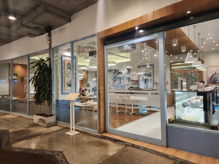 The glass front of Krua Bangna at night, with diners at pale wood tables inside, botanical prints on the walls and a cake counter beside the door.