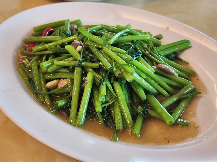 Stir-fried morning glory with garlic cloves and red chillies in a light brown sauce on a white oval plate.