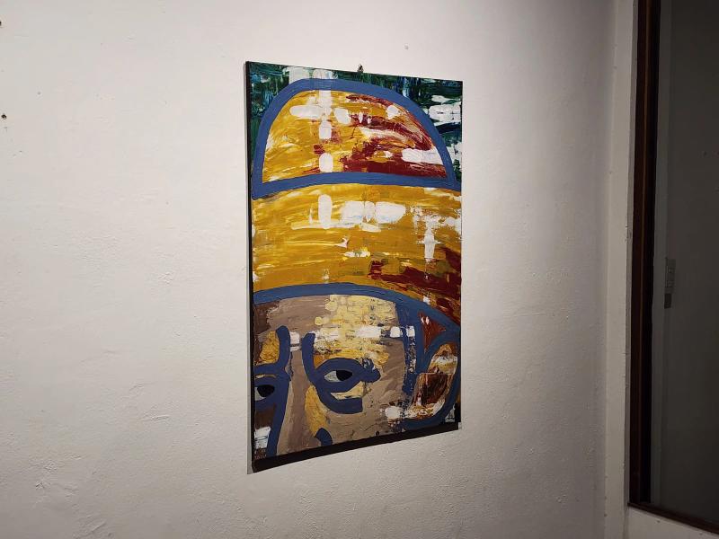 An abstract painting of a face under a yellow hat outlined in blue, hung on a white plaster wall.