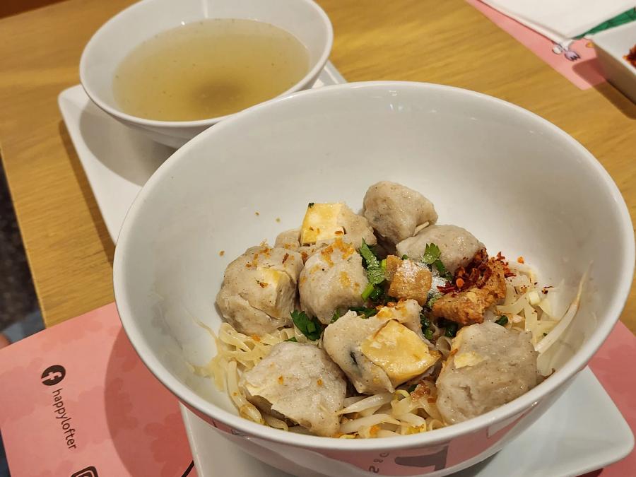 A bowl of dry egg noodles topped with fishballs, stuffed tofu and a crispy fried topping, with a bowl of clear broth behind.