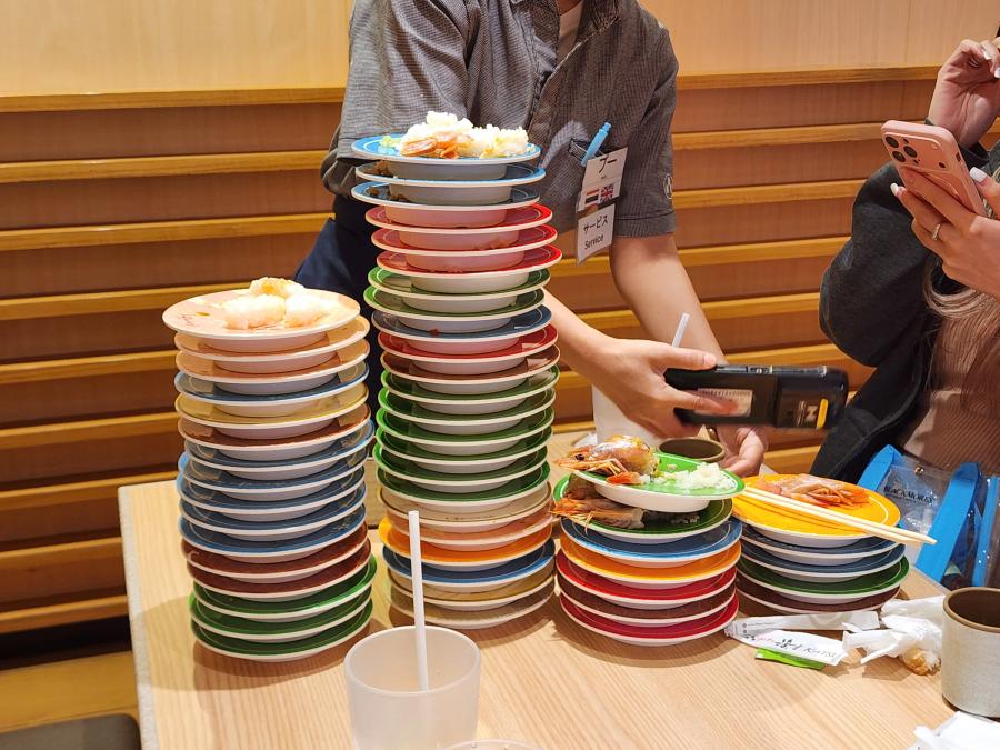 Four towers of empty sushi plates in mixed colours on a wooden table, with prawn heads on the top plates, while a member of staff scans them with a handheld reader.