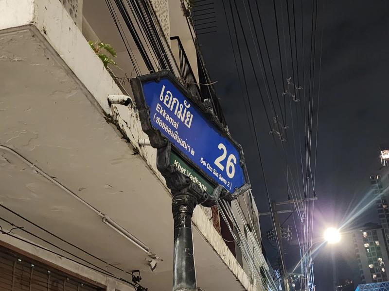 A blue Bangkok street sign reading Ekkamai 26 in Thai and English, photographed from below at night against overhead cables.