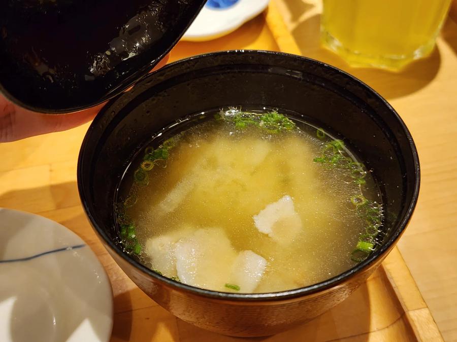 A black lacquer bowl of miso soup with pieces of pork and spring onion, its lid held above it.