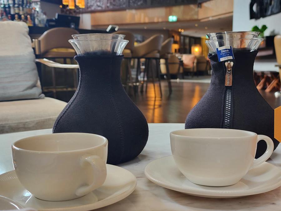 Two glass teapots in black insulated jackets and two empty white cups on a marble table, with the bar stools and lounge of the Diplomat Bar behind.