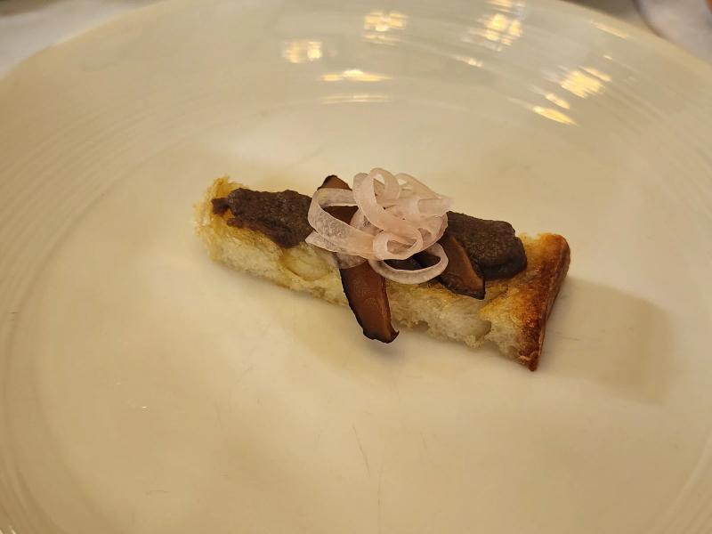 A finger of toast topped with mushroom parfait, slices of dark shiitake and curls of pickled shallot on a white plate.
